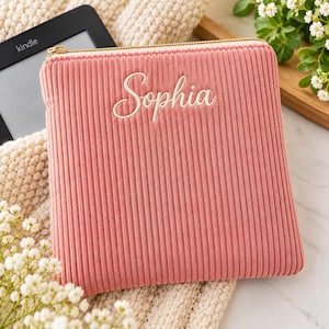 May include: A square, dusty pink corduroy pouch featuring vertical ribbed texture and a gold-toned zipper closure. The name Sophia is embroidered in white cursive script across the center of the front panel of the fabric accessory.