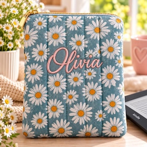 May include: A rectangular, padded protective sleeve for an e-reader or tablet, featuring a blue background with a white daisy pattern. The name Olivia is embroidered in pink script in the center. It has a gold-toned metal zipper closure.
