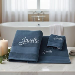 May include: Three navy blue personalised bath towels embroidered with the name "Janelle" in white script. The towels are on a marble surface with lit candles and decorative stones in a bathroom setting.