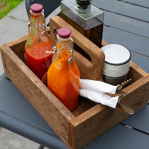 Puede incluir: Una bandeja de servir de madera con dos botellas de vidrio llenas de salsas roja y naranja, un recipiente plateado y servilletas blancas. La bandeja tiene un asa y está sobre una mesa gris.