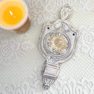 May include: A vintage-style, ornate silver-colored hand mirror with a decorative floral design. The mirror has a shield-like shape with a clear glass center. A lit candle in a glass jar is in the background.