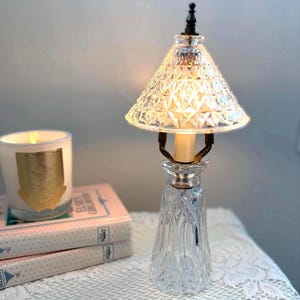 May include: A vintage crystal table lamp with a diamond-patterned shade and a clear, fluted base. The lamp has a brass-colored candle holder and a black finial. A lit candle and a stack of books are in the background.