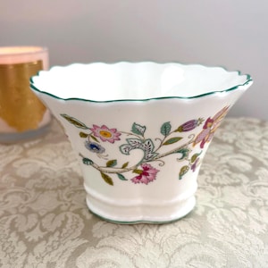 May include: A white porcelain bowl with a scalloped rim and a green painted edge. The exterior features a delicate floral pattern with pink, blue, and purple flowers on green stems. The bowl has a flared shape and a small, rounded base.