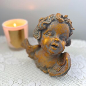 May include: A detailed, antique-style cherub figurine with gold and gray tones. The cherub has curly hair and outstretched wings. A lit candle in a pink glass is in the background, resting on a white lace surface.