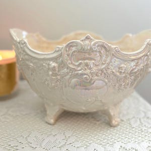 May include: An iridescent white ceramic bowl with ornate embossed detailing. The bowl has a scalloped rim and sits on three small feet. A lit candle is visible in the background.