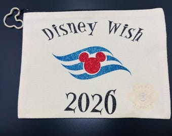 Może przedstawiać: Kremowa kosmetyczka zapinana na zamek z napisem "Disney Wish" nad niebiesko-czerwonym wzorem Myszki Miki i rokiem "2026". Suwak zamka to sylwetka Myszki Miki.