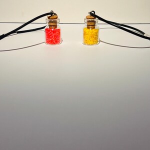 May include: Two glass bottle pendants on black cords. One bottle is filled with small, bright red beads and sealed with a cork stopper. The other bottle is filled with small, bright yellow beads and also sealed with a cork stopper.