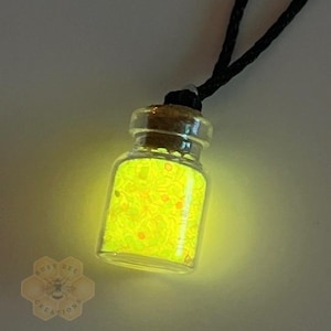 May include: A small glass bottle pendant filled with glowing yellow glitter and small orange specks, suspended from a black cord. The bottle has a cork stopper. The image is lit with a soft, warm glow.