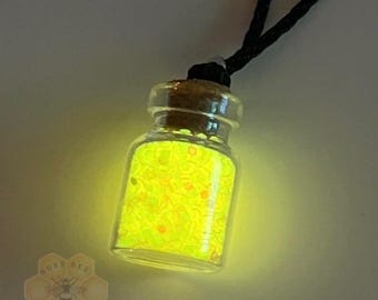 May include: A small glass bottle pendant filled with glowing yellow particles, sealed with a cork stopper, and attached to a black cord. The bottle emits a bright light and features a logo with the text Busy Bee Creations in the bottom corner.