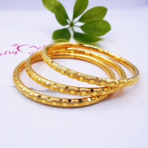 May include: Three gold bangles with a textured surface. The bangles are stacked on top of each other.