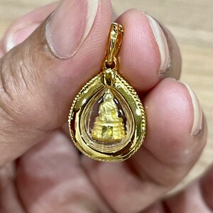 K&ouml;nnte beinhalten: Ein goldener Anh&auml;nger mit einer kleinen goldenen Buddha-Statue in einer durchsichtigen Glaskuppel. Der Anh&auml;nger hat die Form einer Tr&auml;ne und ist mit einem zarten Seil-Muster am Rand verziert.