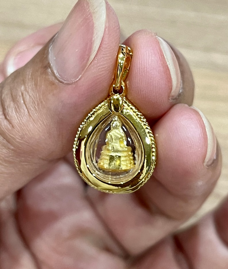 K&ouml;nnte beinhalten: Ein goldener Anh&auml;nger mit einer kleinen goldenen Buddha-Statue in einer durchsichtigen Glaskuppel. Der Anh&auml;nger hat die Form einer Tr&auml;ne und ist mit einem zarten Seil-Muster am Rand verziert.