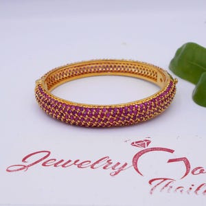 May include: A gold hinged bangle bracelet featuring a dense pavé setting of small, vibrant pink stones covering the outer surface. The interior of the band is smooth gold, and the bracelet includes a secure clasp mechanism on one side.