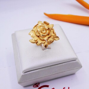 May include: A gold-colored ring with a rose design. The ring is displayed on a white box.