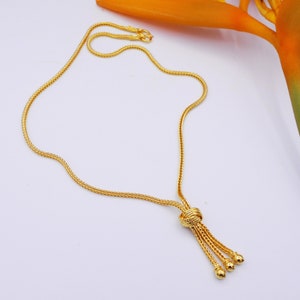 Könnte beinhalten: Eine goldene Kettenkette mit einem geknoteten Anhänger und drei kleinen runden Perlen am Ende. Die Kette ist in einem zarten, geflochtenen Design gehalten.