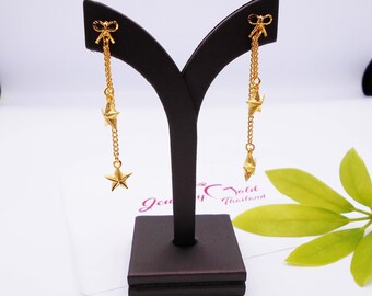 May include: A pair of gold dangle earrings with a star and bow charm. The earrings are displayed on a black stand.