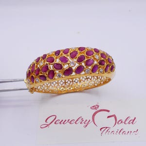 May include: A gold bangle bracelet featuring a cluster of oval-shaped deep pink rubies and small clear round stones. The band has an intricate openwork lattice design. A white card with the text Jewelry Gold Thailand is visible below.