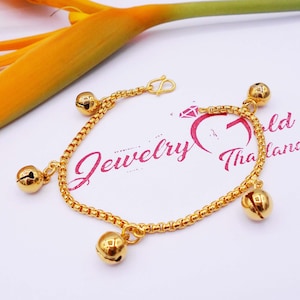 May include: A gold chain bracelet with five small bell charms. The bracelet is on a white surface with a pink and white floral background.