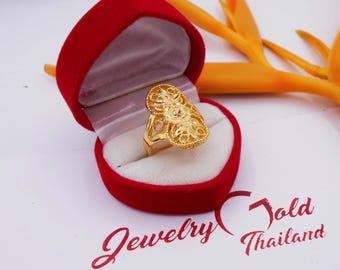 May include: A gold-colored ring with an ornate filigree design, displayed in a red heart-shaped jewelry box. The ring has a detailed, openwork pattern. The box is lined with white fabric. The background includes a yellow flower and the text "Jewelry Gold Thailand."