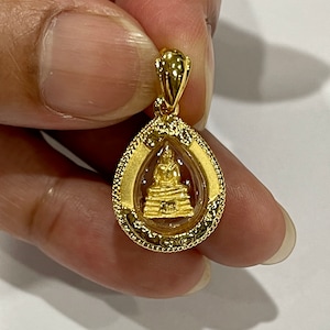 K&ouml;nnte beinhalten: Ein goldener Anh&auml;nger mit einer kleinen Buddha-Statue in einem transparenten Glasoval. Der Anh&auml;nger hat einen detaillierten goldenen Rand und eine Schlaufe zum Anbringen einer Kette.