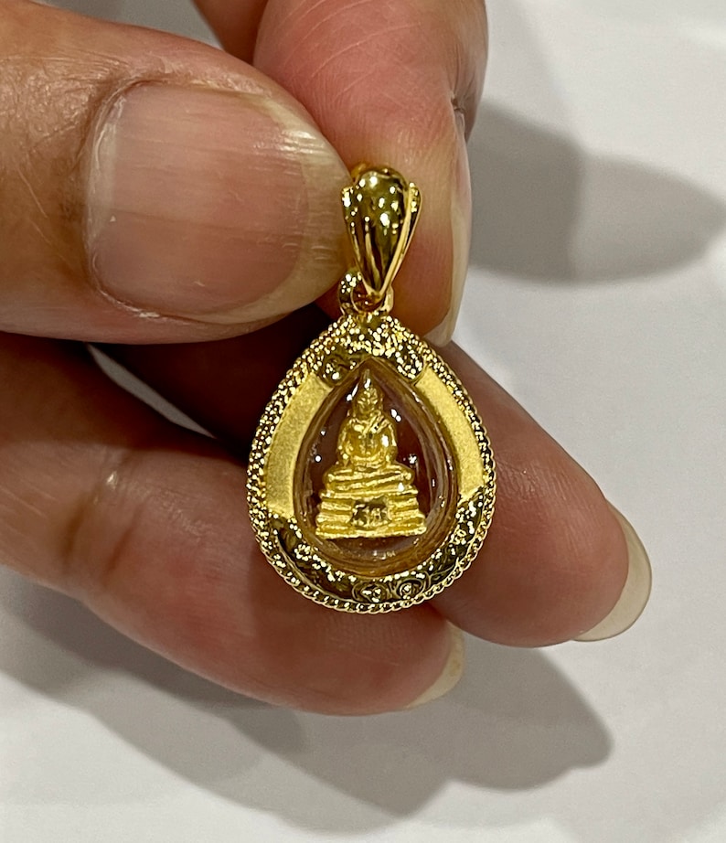 K&ouml;nnte beinhalten: Ein goldener Anh&auml;nger mit einer kleinen Buddha-Statue in einem transparenten Glasoval. Der Anh&auml;nger hat einen detaillierten goldenen Rand und eine Schlaufe zum Anbringen einer Kette.