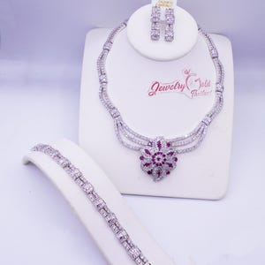 May include: A jewelry set consisting of a necklace, a bracelet, and earrings. The necklace features three silver-toned chains leading to a central floral pendant with red and clear stones. The bracelet and earrings match the necklace design.