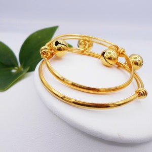 May include: Two gold bangles with bells, one slightly larger than the other, on a white display stand.
