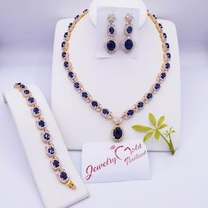 Könnte beinhalten: Ein goldfarbenes Schmuckset mit Halskette, Armband und Ohrringen. Die Stücke sind mit ovalen, dunkelblauen Edelsteinen und klaren Akzentsteinen verziert. Das Set wird auf weißen Ständern präsentiert, mit einer Visitenkarte "Jewelry Gold Thailand".
