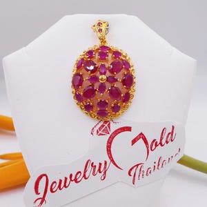 May include: A gold pendant with a round design featuring a cluster of red ruby stones. The pendant is on a white display stand with the text "Jewelry Gold Thailand".