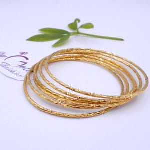 May include: A set of six gold-colored bangles with a textured surface. The bangles are stacked together on a white surface, with a green leaf and a tag with text in the background. The bangles are a bright, shiny gold color.
