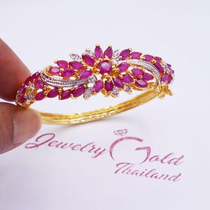 May include: A gold-toned bracelet with a floral design. The bracelet features numerous oval-shaped, deep pink gemstones and small, clear accent stones. The bracelet is held against a white background with the text "Jewelry Gold Thailand".