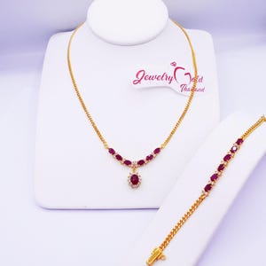 May include: A gold necklace and bracelet set featuring oval-cut red gemstones and small clear stones. The necklace has a pendant with a large red gemstone surrounded by clear stones. The bracelet has a matching design. The jewelry is displayed on white stands.