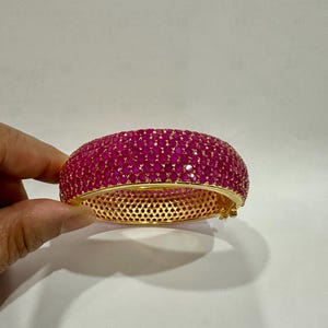 May include: A gold-toned bangle bracelet encrusted with numerous small, oval-shaped, magenta-colored gemstones. The bracelet has a perforated design on the inner band and is held against a white background.
