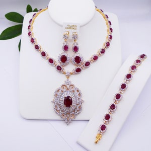 May include: A gold-tone necklace, bracelet, and earrings set with red oval stones and clear cubic zirconia accents. The pendant features a large red oval stone surrounded by clear cubic zirconia stones.