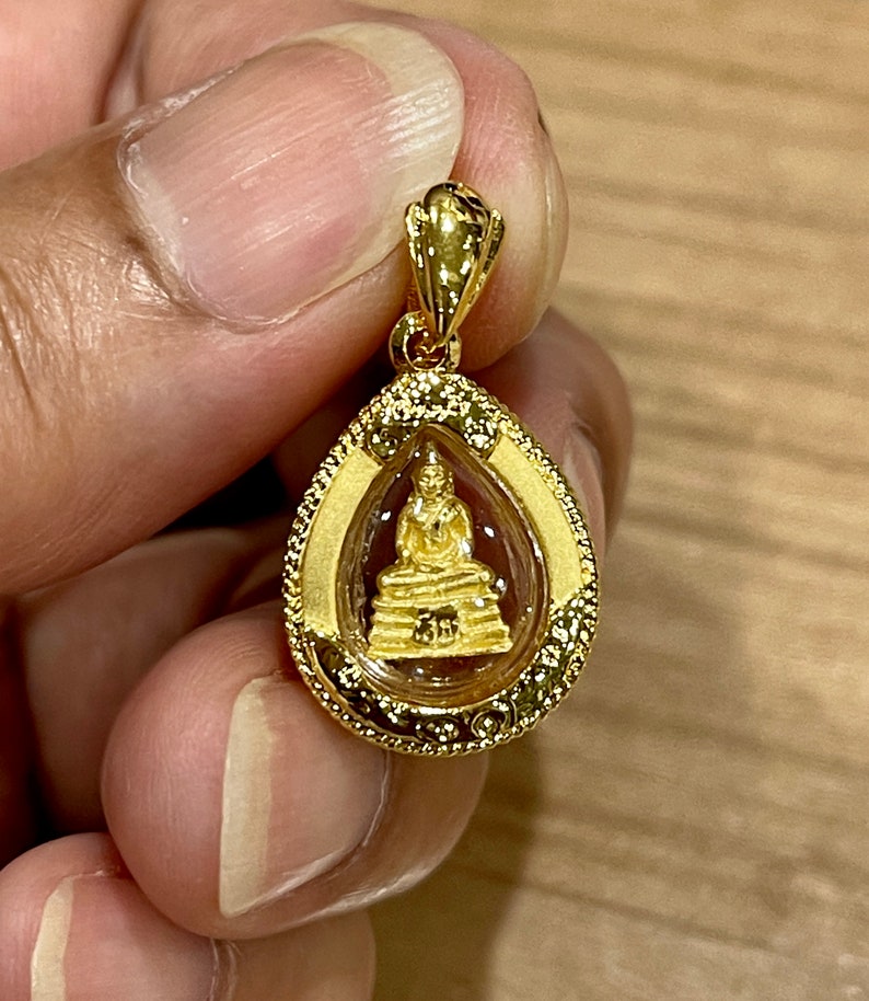 K&ouml;nnte beinhalten: Ein goldener Anh&auml;nger mit einem kleinen, detaillierten Bild eines sitzenden Buddha in meditativer Pose. Der Anh&auml;nger ist tropfenf&ouml;rmig und hat einen dekorativen Rand.
