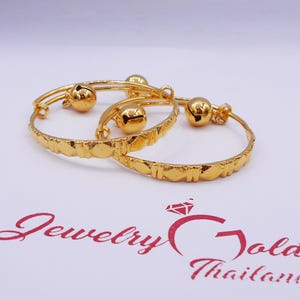 Könnte beinhalten: Zwei goldene Babyarmbänder mit Glöckchen. Die Armbänder sind mit einem strukturierten Muster verziert. Die Armbänder sind übereinander gestapelt.