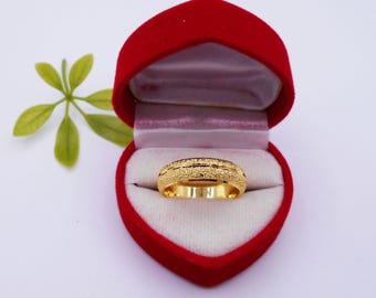 May include: A gold ring with a textured surface sits inside a red heart-shaped box. The ring has a band with a detailed pattern. The box is open, revealing a white interior. A green leafy sprig is in the background.