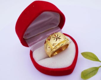 May include: A gold ring with a starburst design is displayed inside a red heart-shaped box. The ring has a textured surface and is set against a white interior. The box is open, showcasing the ring's intricate details.