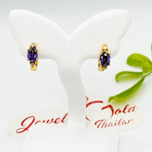 May include: A pair of gold hoop earrings, each featuring a large oval purple gemstone and small clear accent stones. The earrings are displayed on a white stand against a white background.