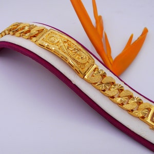 May include: A gold bracelet with a detailed design featuring a dragon. The bracelet is on a white and purple display stand.