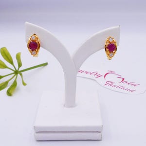 May include: A pair of gold hoop earrings with a red gemstone and small white gemstones. The earrings are on a white display stand.