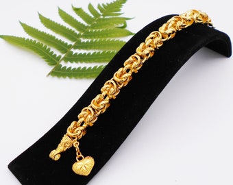 Könnte beinhalten: Ein goldfarbenes Armband mit einem herzförmigen Anhänger. Das Armband besteht aus einer dicken, geflochtenen Kette.
