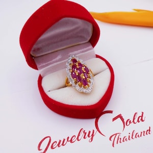 May include: A gold ring featuring a cluster of oval-cut, deep red gemstones encircled by a halo of small, clear stones. The ring is presented in an open, red heart-shaped jewellery box. The box is on a white surface.