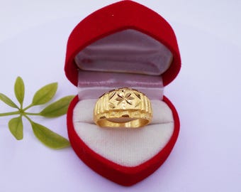 May include: A gold ring with a starburst design is displayed inside a red heart-shaped box. The ring has a textured band and a detailed top. The box is open, revealing a white interior.