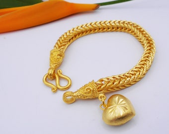 Könnte beinhalten: Ein goldenes Armband mit einem herzförmigen Anhänger. Das Armband besteht aus einer dicken, geflochtenen Kette mit aufwendigen Details. Der Herzanhänger ist ebenfalls golden und hat eine strukturierte Oberfläche.