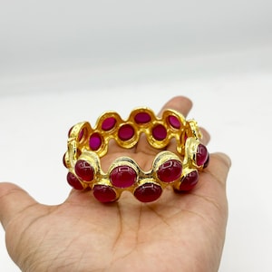 May include: A gold-colored bangle bracelet with oval, ruby-red stones. The bracelet has a repeating pattern of the red stones set in a gold-tone metal. The bracelet is shown on a light background.