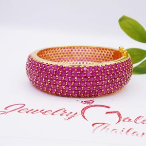 May include: A gold-toned bangle bracelet encrusted with numerous small, vibrant magenta-colored gemstones. The bracelet has a rounded shape and a secure clasp. The background is white, with a sprig of green leaves in the upper right corner.