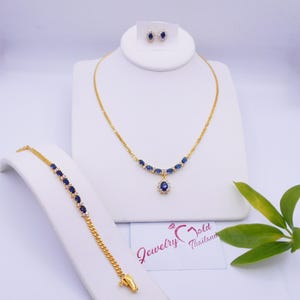 May include: A gold-coloured jewellery set comprising a necklace, bracelet, and earrings. The necklace and bracelet feature blue oval stones. The earrings have a single blue stone. The jewellery is displayed on white stands.