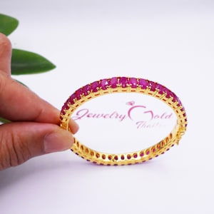 May include: A gold bangle bracelet with a row of pink gemstones around the entire circumference. The bracelet is on a white surface.