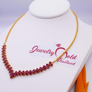 May include: A gold chain necklace with a ruby stone pendant. The necklace is on a white display stand. The text "Jewelry Gold Thailand" is visible on the stand.